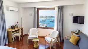 A hotel room with a bed, a desk, two chairs, a small table, a sofa, a TV, and a window with a view of boats and water. - Best Western Hotel Martello (Lampedusa)