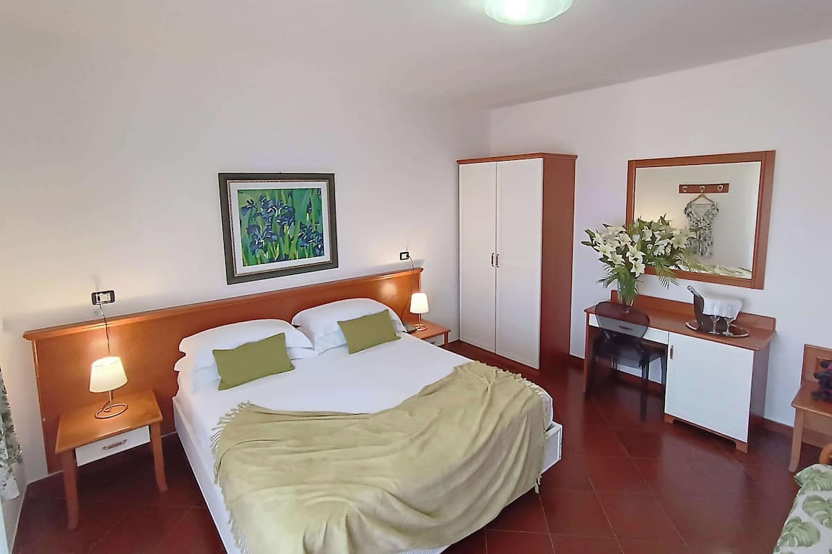Ein Hotelzimmer mit Bett, Nachttischen, einem Kleiderschrank, einem Schreibtisch mit Stuhl, einem Spiegel und einem gerahmten Bild an der Wand.