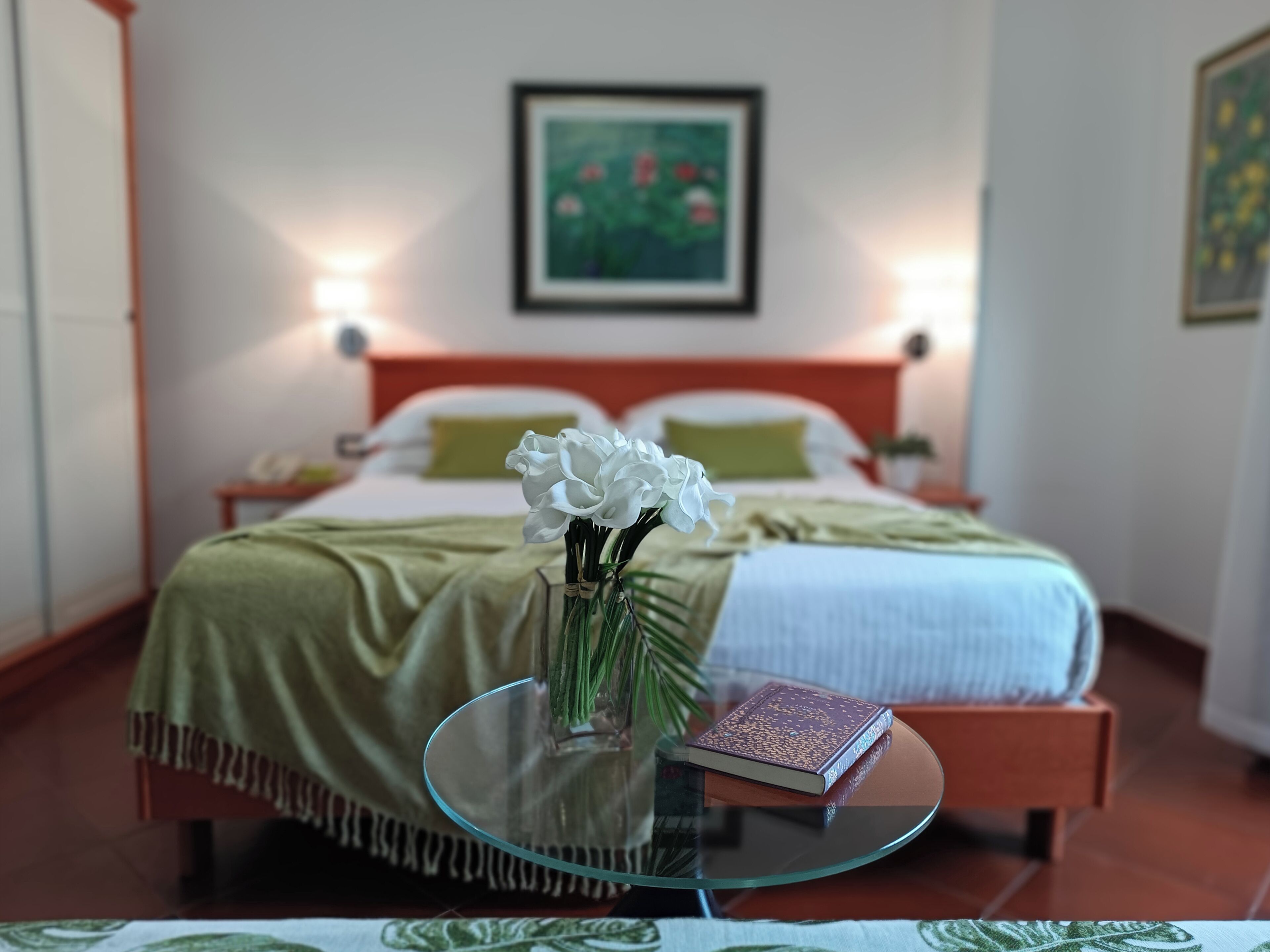 A neatly made bed with a green blanket, a bedside table with a book, and a vase with white flowers.