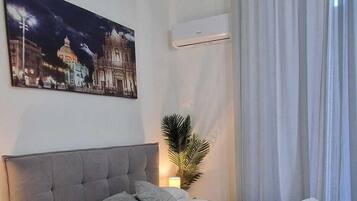 A bedroom with a large bed, a night sky cityscape painting, a potted plant, and a window with curtains.