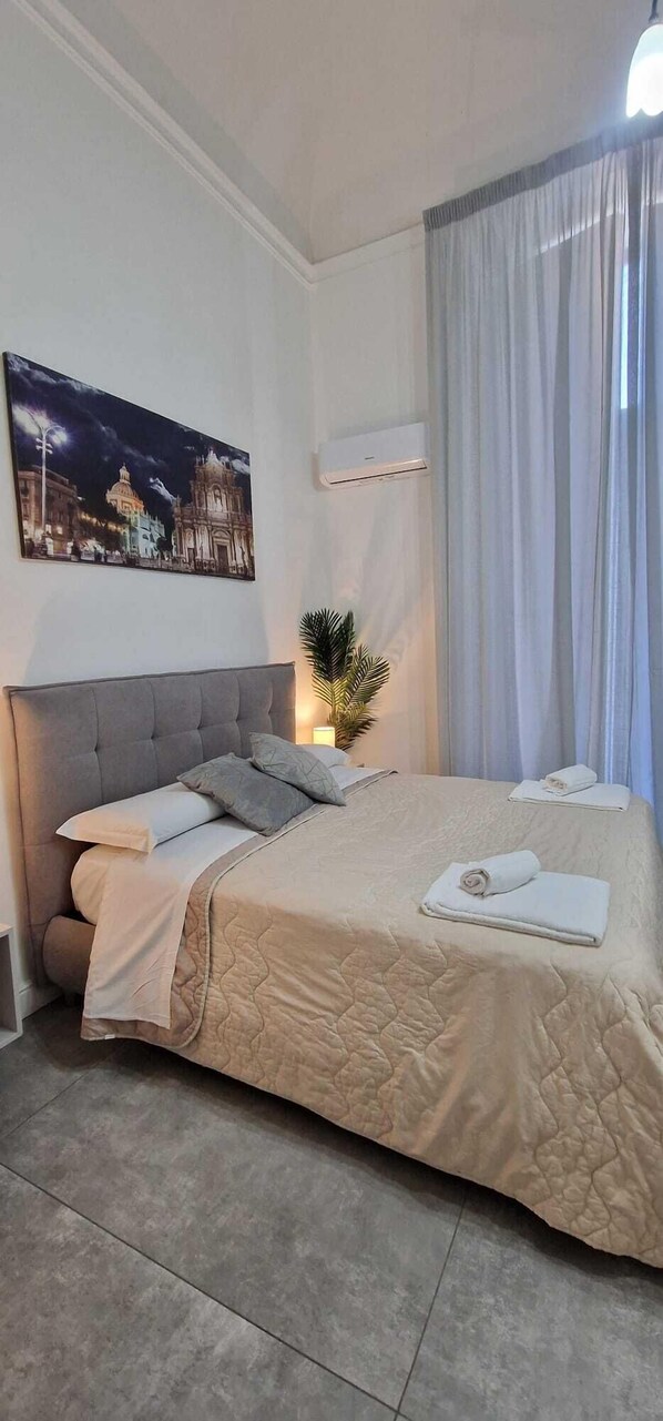 A bedroom with a large bed, a night sky cityscape painting, a potted plant, and a window with curtains.