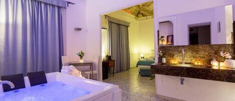 Habitación de hotel con una bañera grande, una zona de estar con sofá, una mesa de comedor y un área de cocina con mostrador.