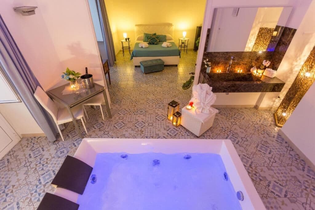 A hotel room with a large bathtub, a bed with green bedding, a small table with a plant, and a bathroom with a marble countertop.
