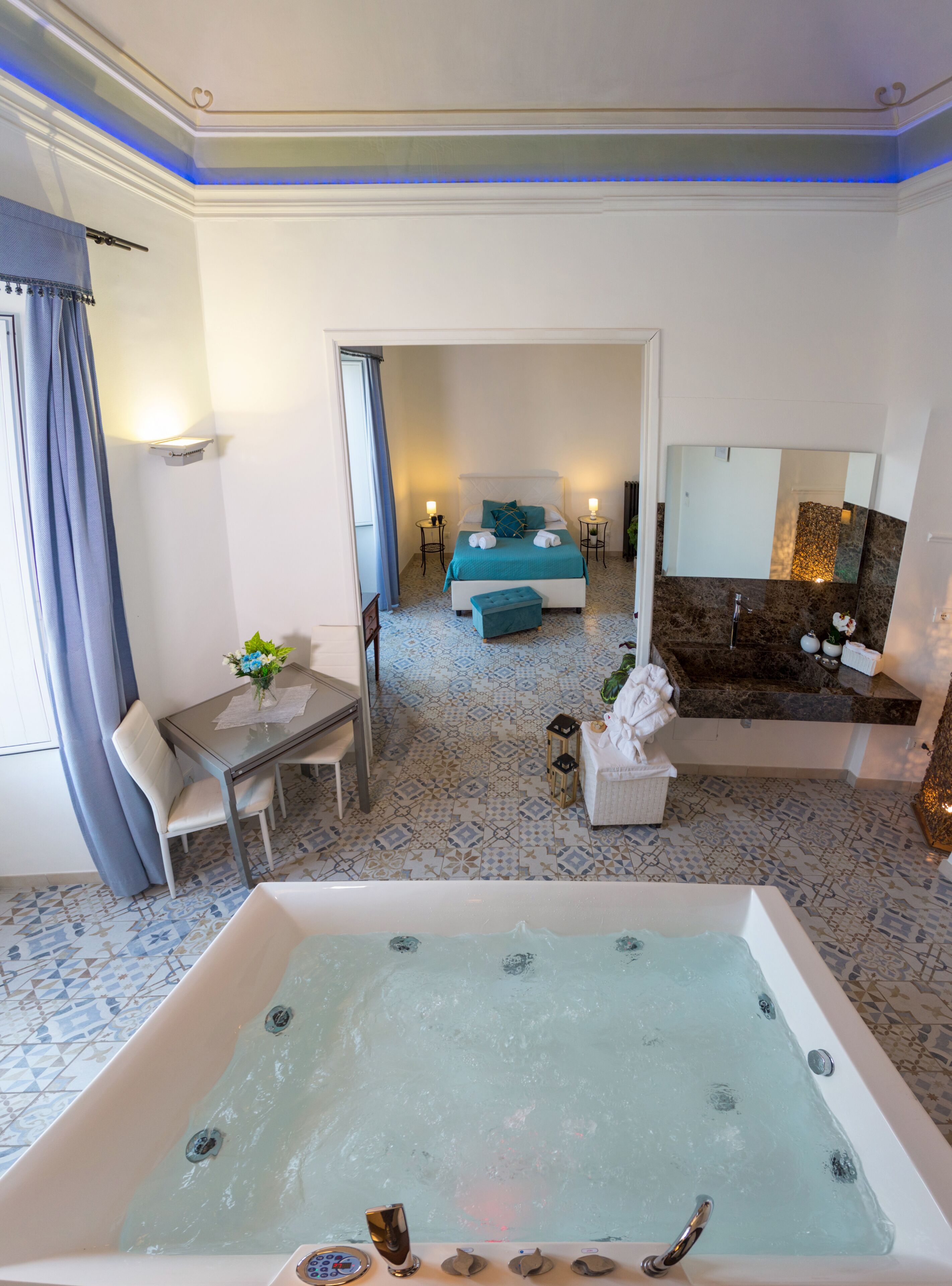 A hotel room with a large bathtub, a bed with a blue headboard, a small table with a vase, and a bathroom with a marble countertop and a mirror.
