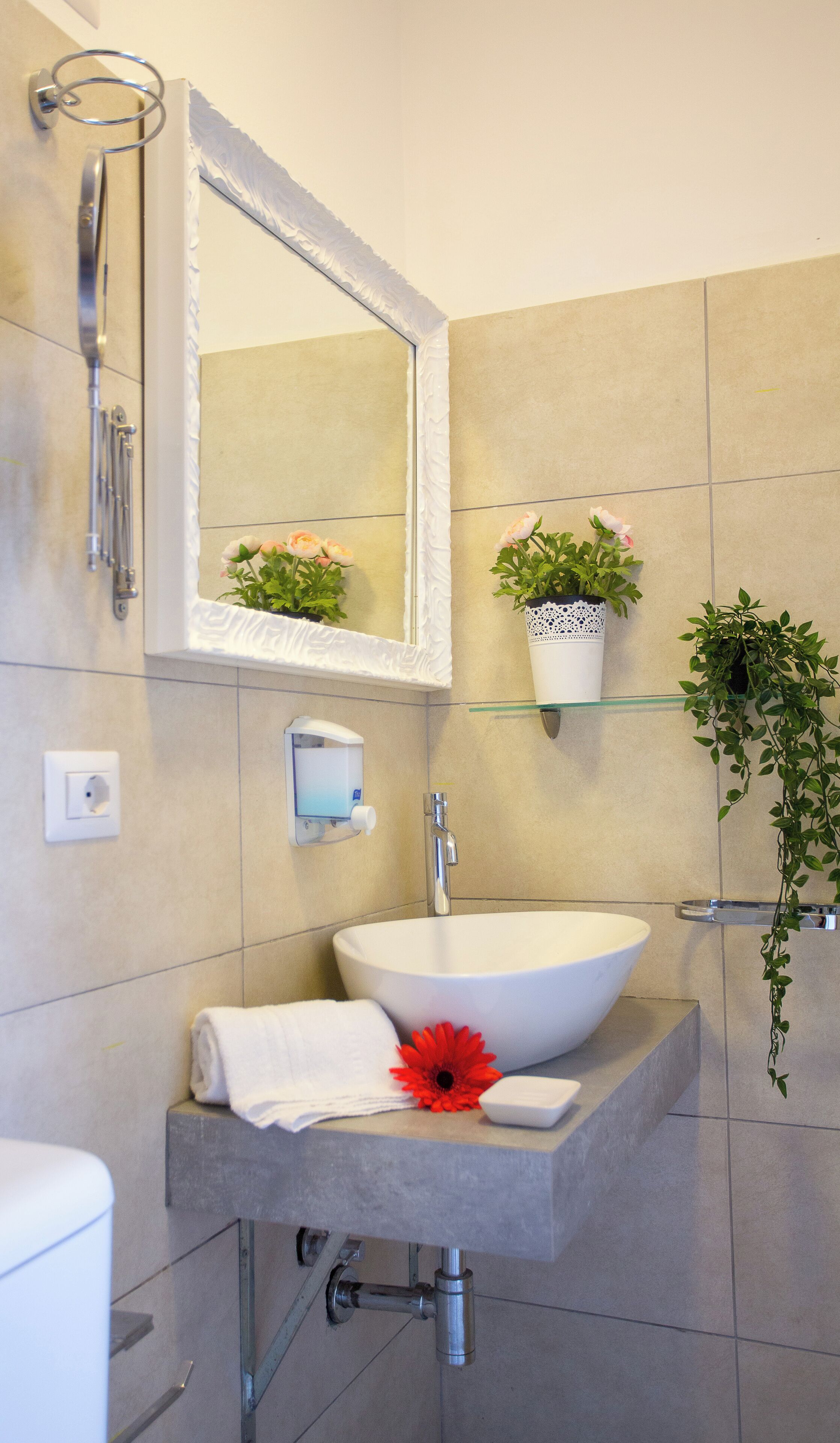 A modern bathroom with a white sink, a large mirror, and a shower. There are potted plants on the wall and a towel on the counter.