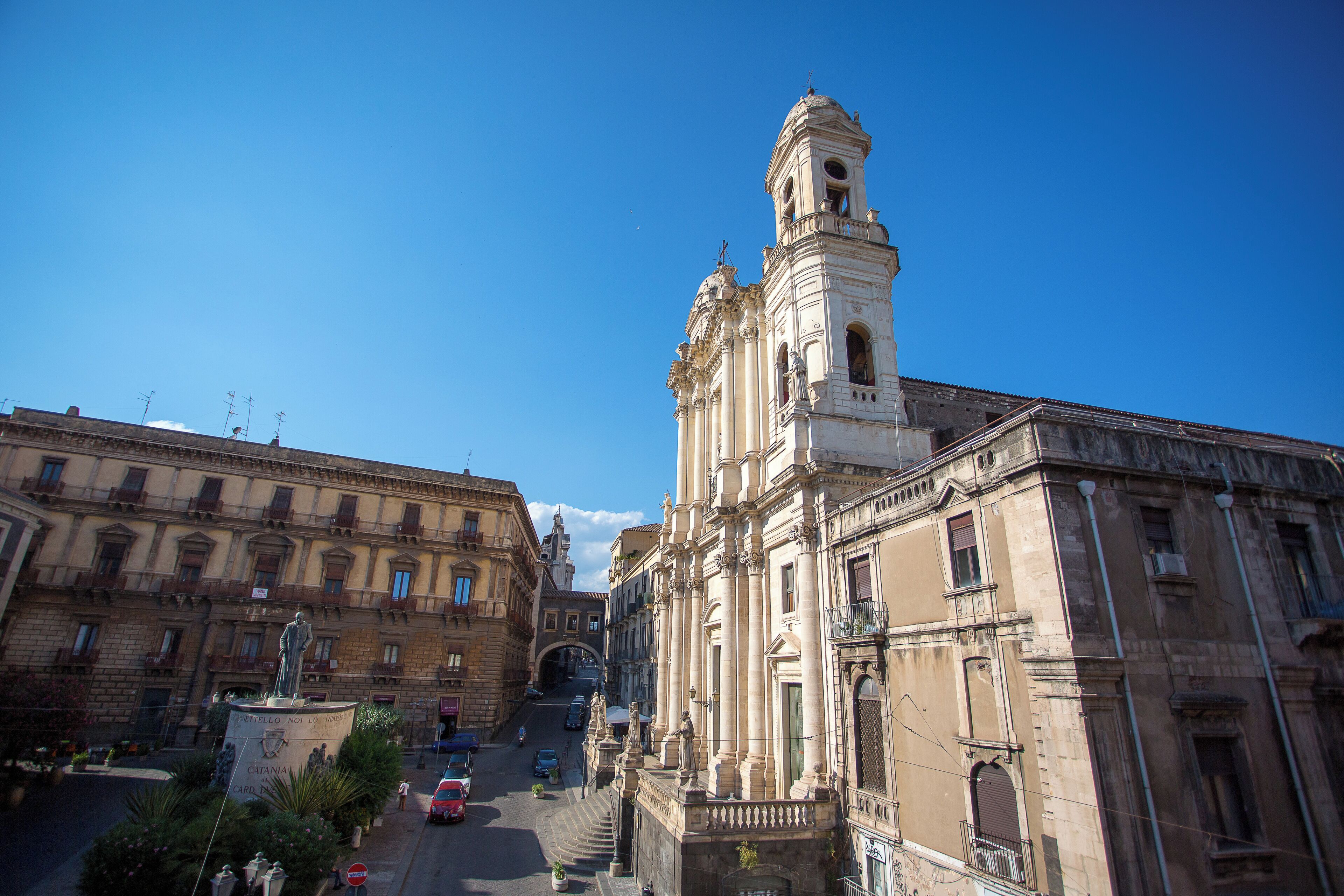 A historic building with a bell tower, a statue in front, and a street with cars and pedestrians.