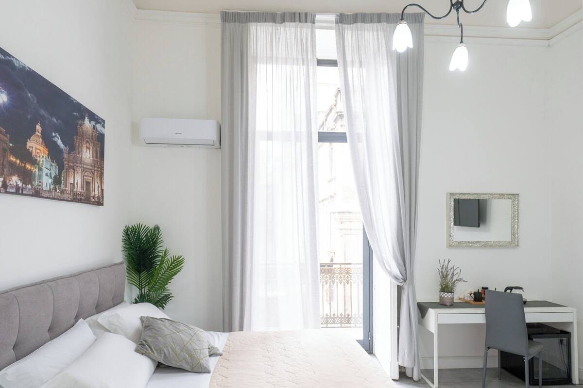 A bedroom with a bed, a desk, and a chair. There is a window with curtains, a plant, and a painting on the wall.