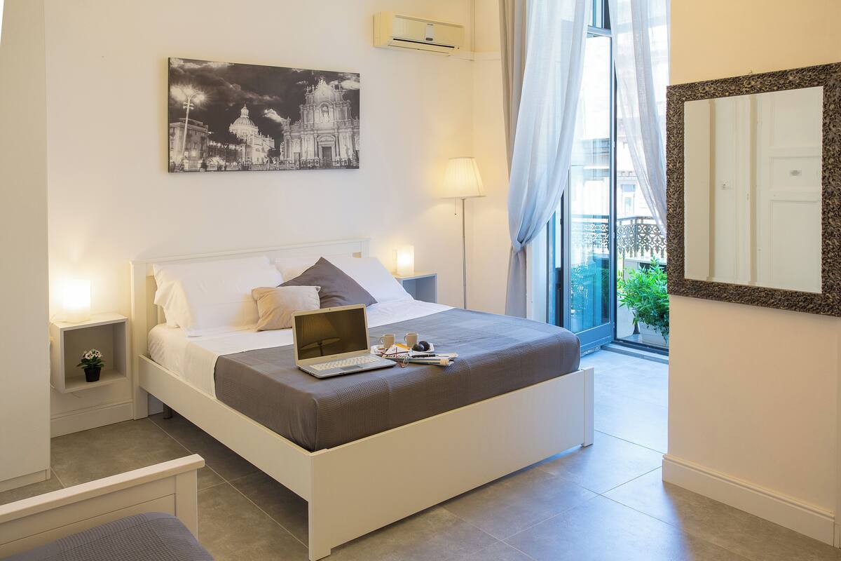 A modern bedroom with a bed, a laptop, a bedside table with a lamp, and a cityscape painting on the wall.