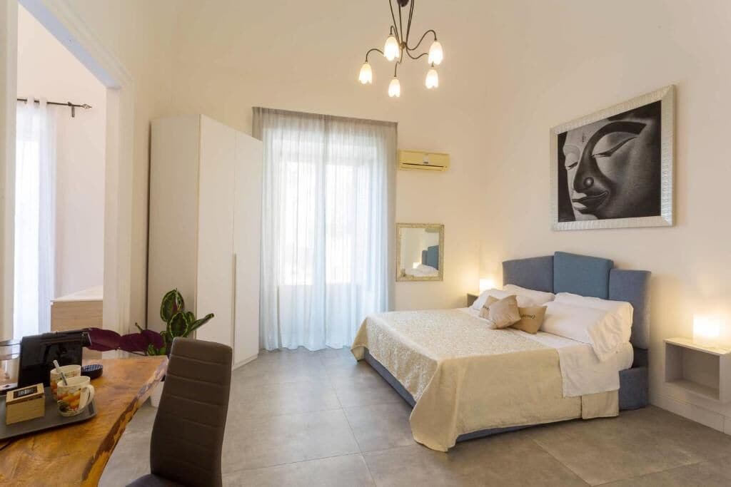 A bedroom with a bed, a desk, a chair, a plant, a painting, and a window with curtains.