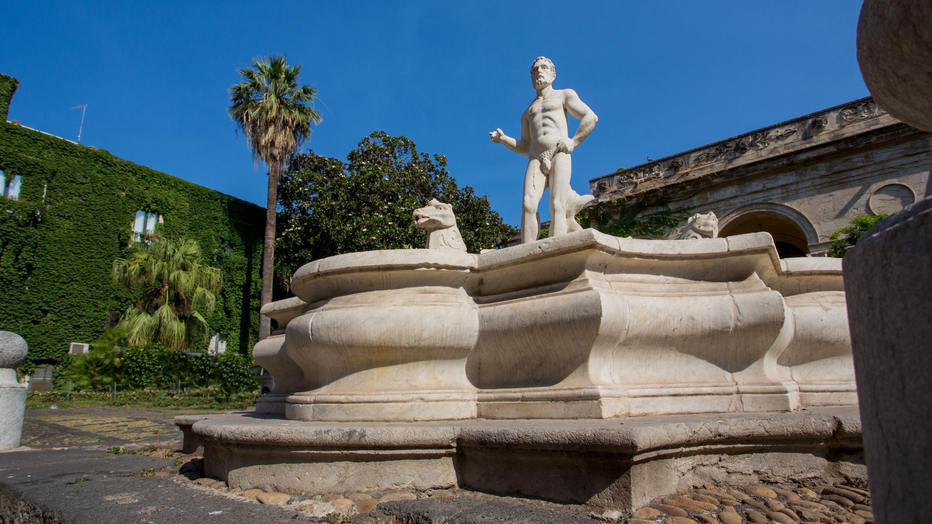 A classical fountain with a statue, surrounded by greenery and a building with classical architecture.
