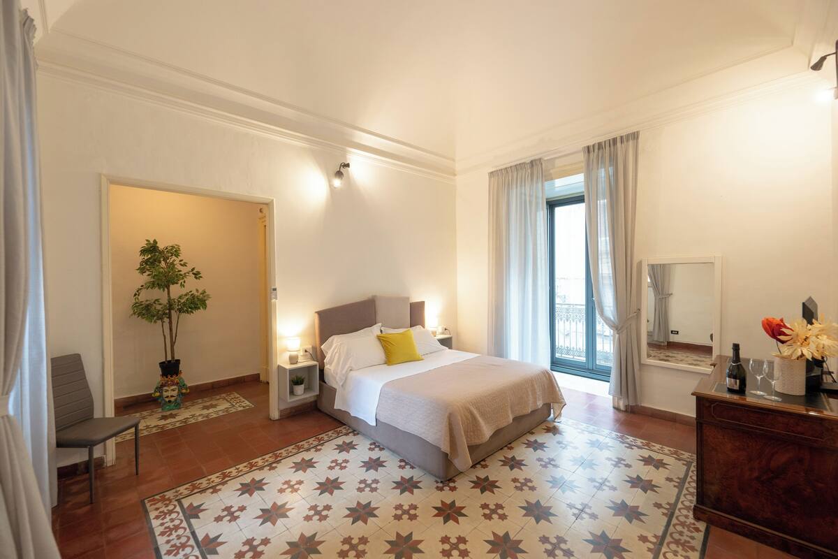 A hotel room with a bed, bedside tables, a nightstand, a potted plant, a dresser, and a window with curtains.