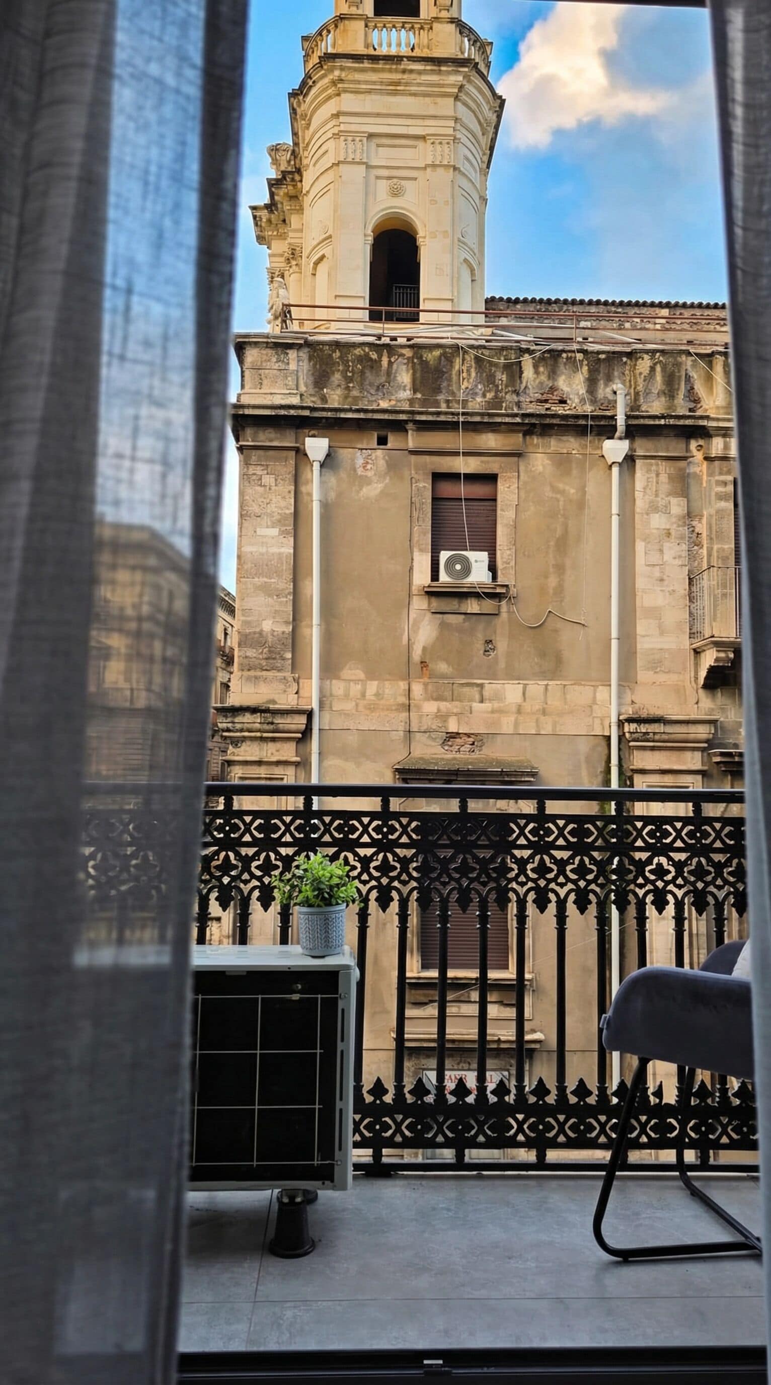 A balcony with a view of a historic building with a bell tower, a potted plant, and an air conditioning unit.