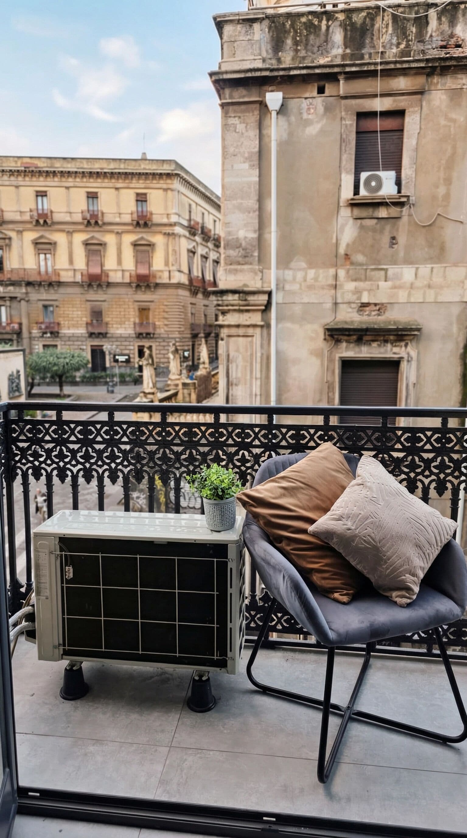 A balcony with a black metal railing, a grey cushioned chair with a plant and cushions, and an air conditioning unit.
