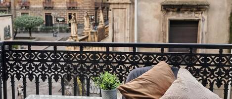 A balcony with a black metal railing, a grey cushioned chair with a plant and cushions, and an air conditioning unit.