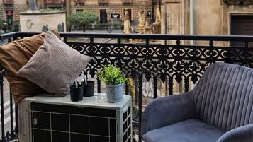 Un balcón con una silla gris, una mesita con una planta y un cojín beige. Al fondo, un edificio de arquitectura clásica y estatuas.