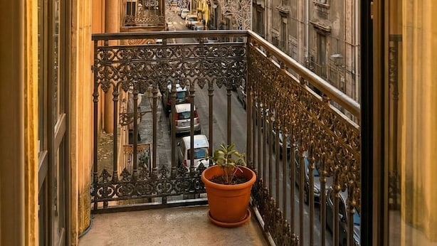 Un balcón con barandillas de hierro ornamentadas, una planta en maceta y vistas de un paisaje urbano con edificios históricos y una estructura en forma de cúpula.