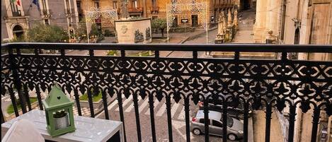Un balcón con una silla y una mesa, con vistas a una calle con edificios históricos y una fuente.