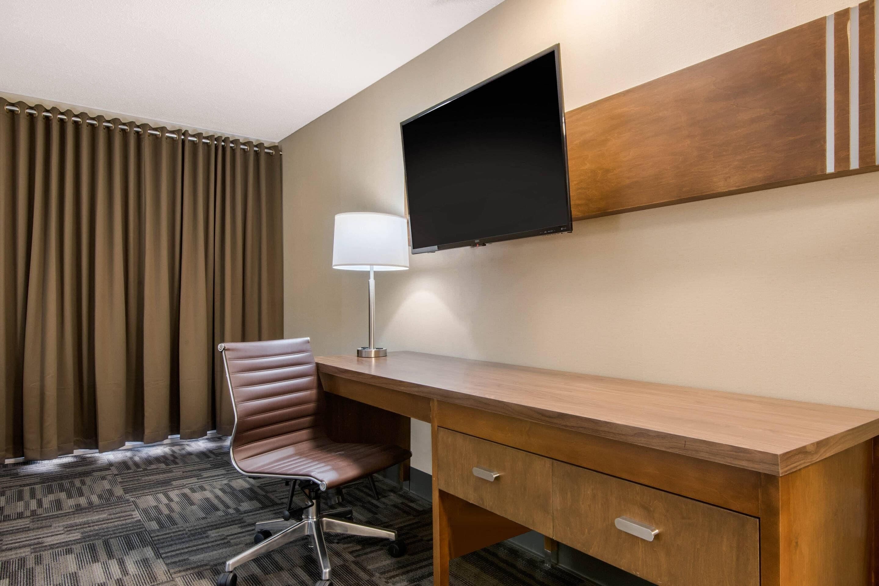 Habitación de hotel con escritorio de madera, sillón de cuero, televisor de pantalla plana en la pared, lámpara y alfombra estampada.