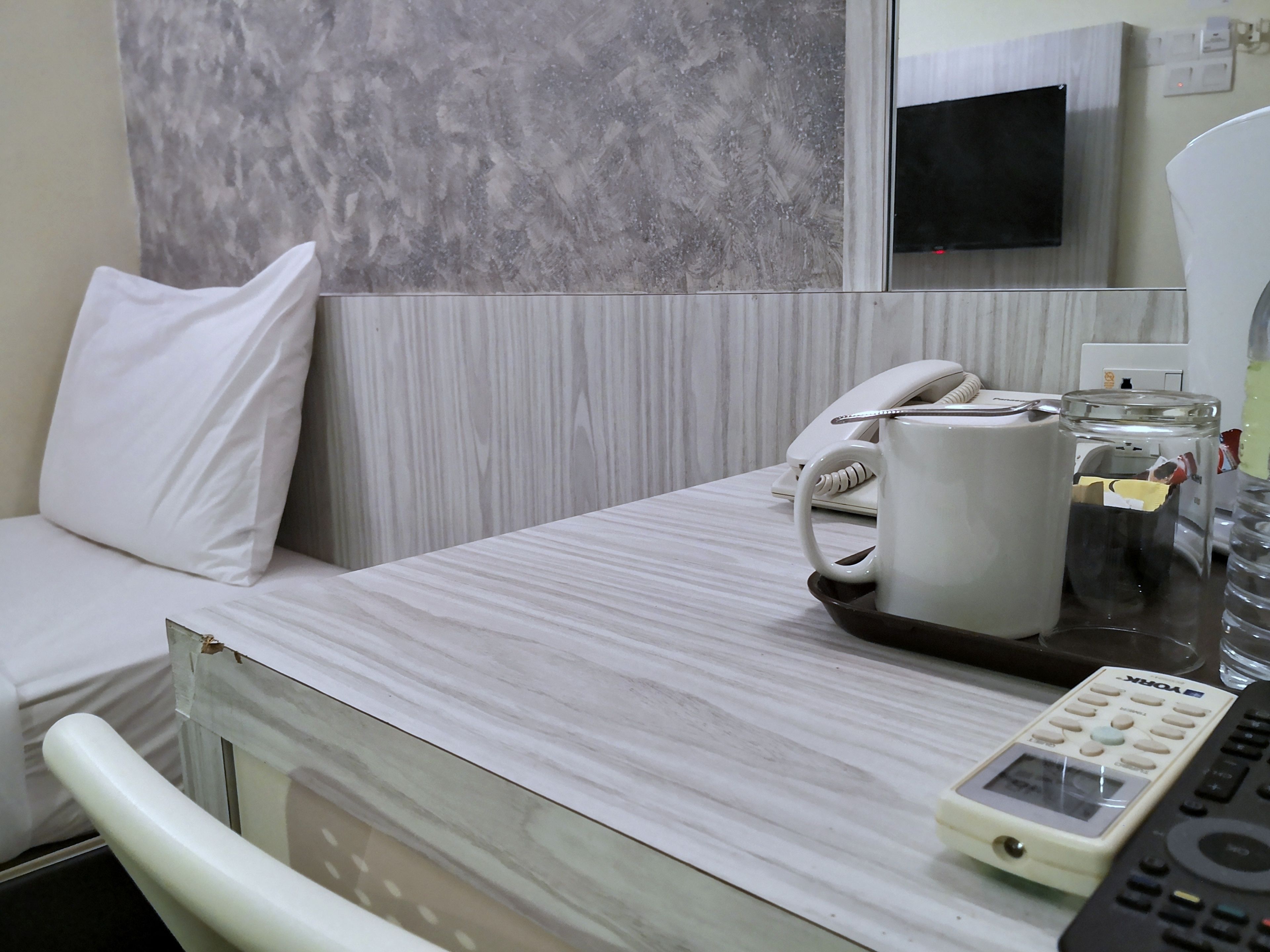 A hotel room with a bed, a bedside table with a telephone, a glass jar, and a remote control.
