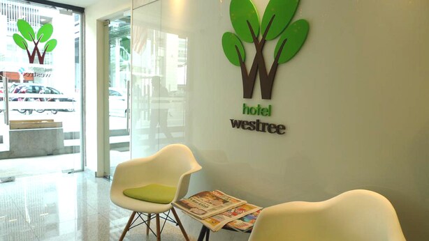 A hotel reception area with a logo, two white chairs with green cushions, and a newspaper on a small table. - Hotel Westree (Kuala Lumpur)