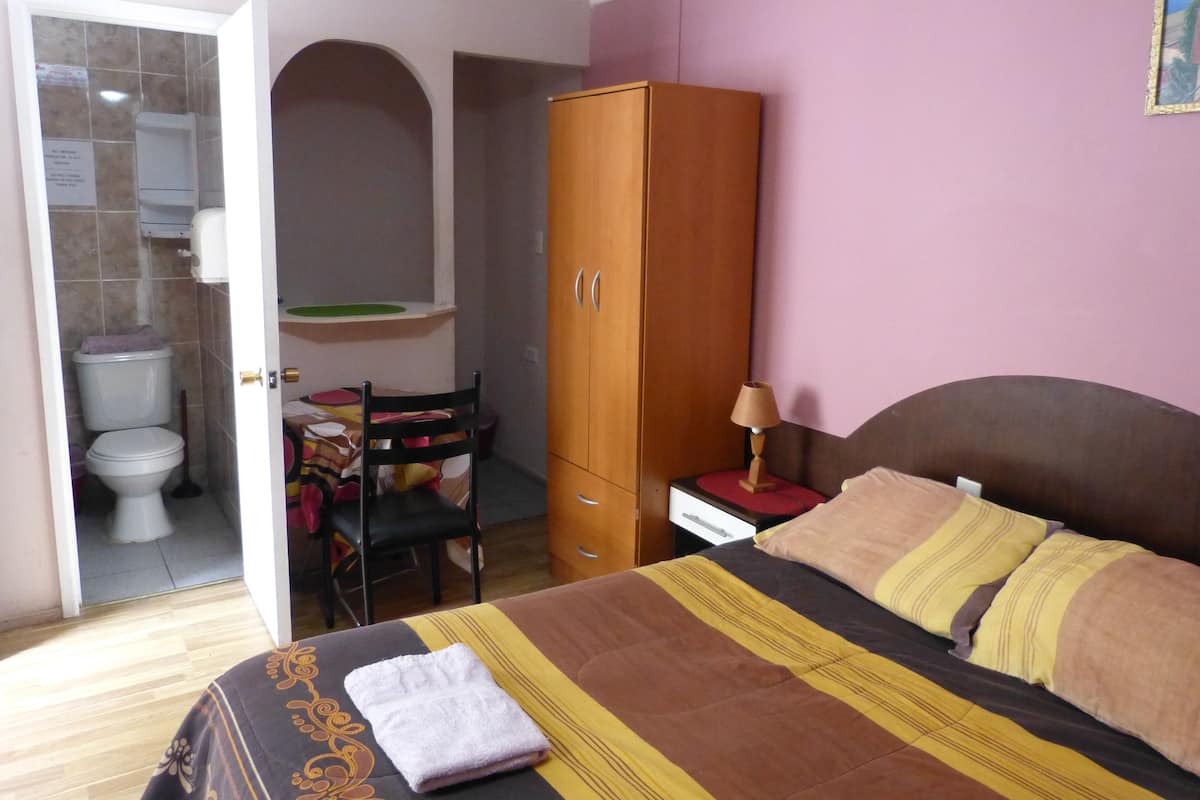 A bedroom with a bed, a wooden wardrobe, a chair, and a small table with a lamp.