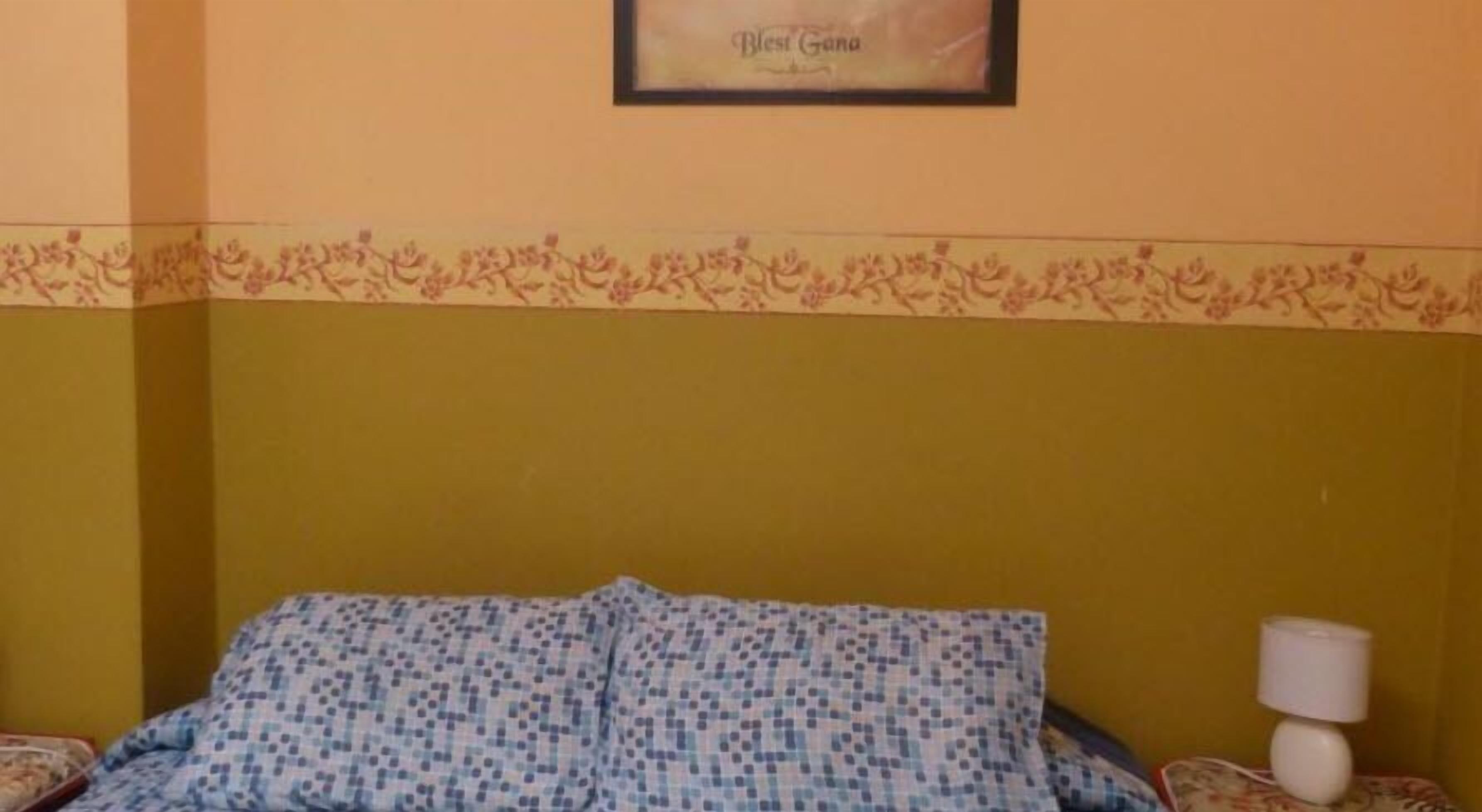 A room with a bed covered in a patterned blanket, a wall with a floral border, and a framed item above the bed.