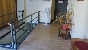 A hallway with a wooden floor, a metal railing, a fan, and a potted plant with yellow flowers.