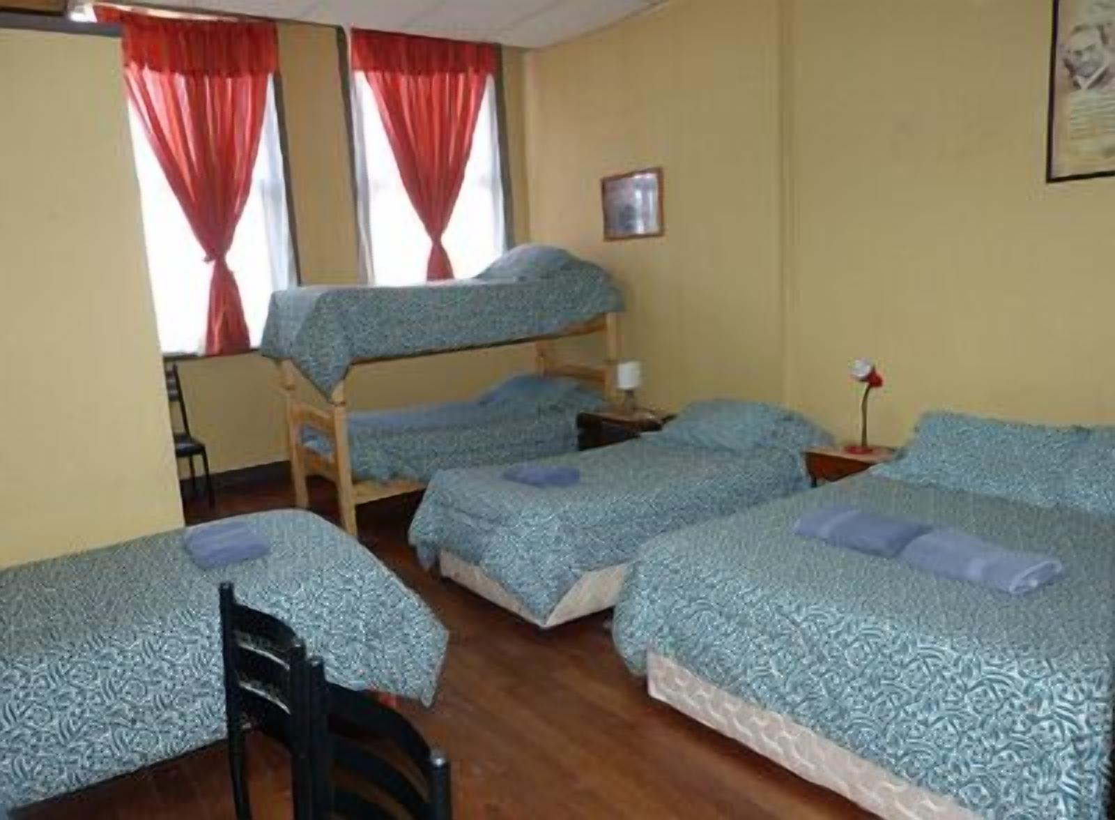 A dormitory room with bunk beds, a chair, a bedside table, and a window with red curtains.