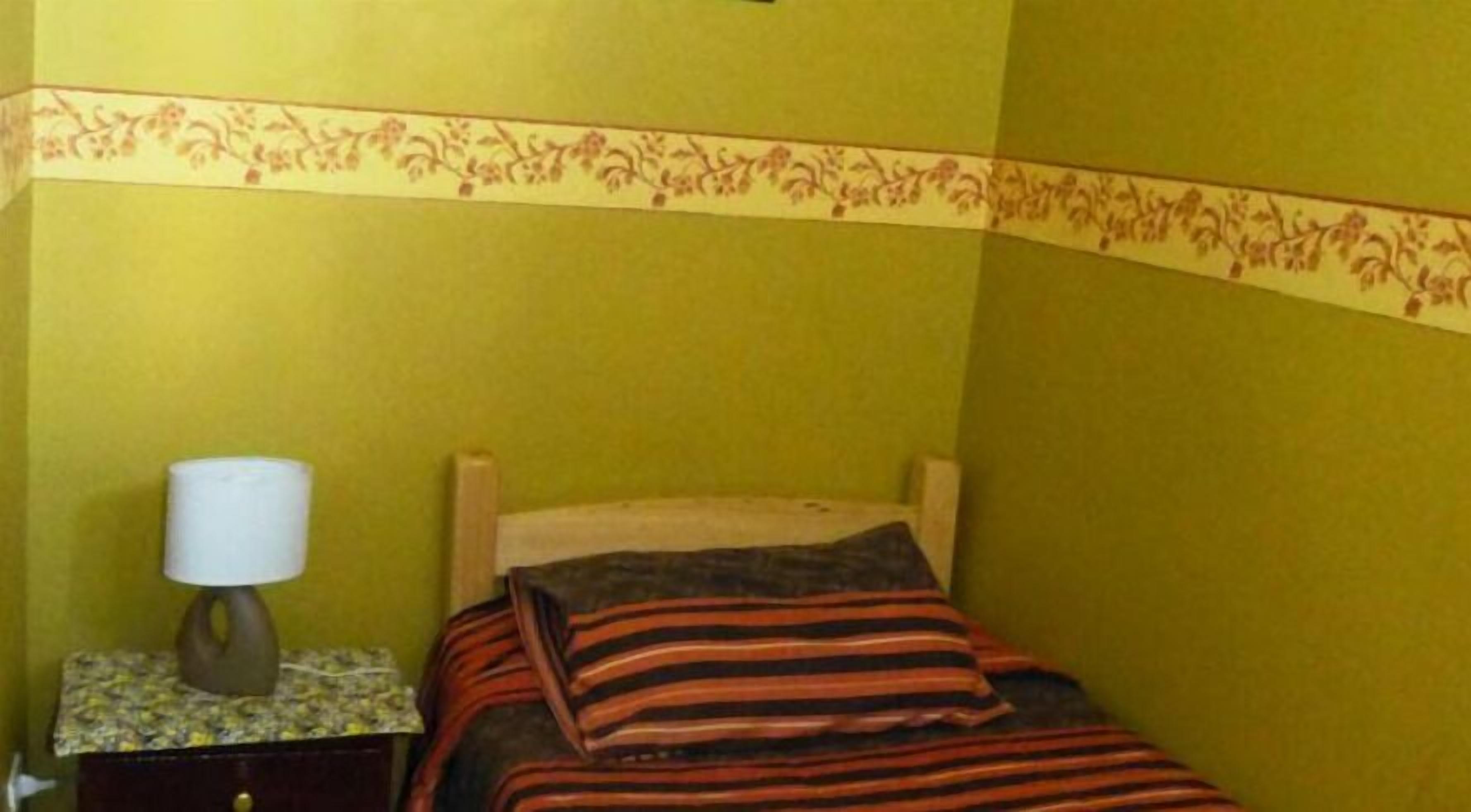 A small, single-bed room with a wooden headboard, a floral-patterned border above the bed, and a bedside table with a lamp.