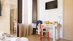 Habitación de hotel con cama, escritorio con silla, televisor y una pequeña mesa con frutas y un cactus.