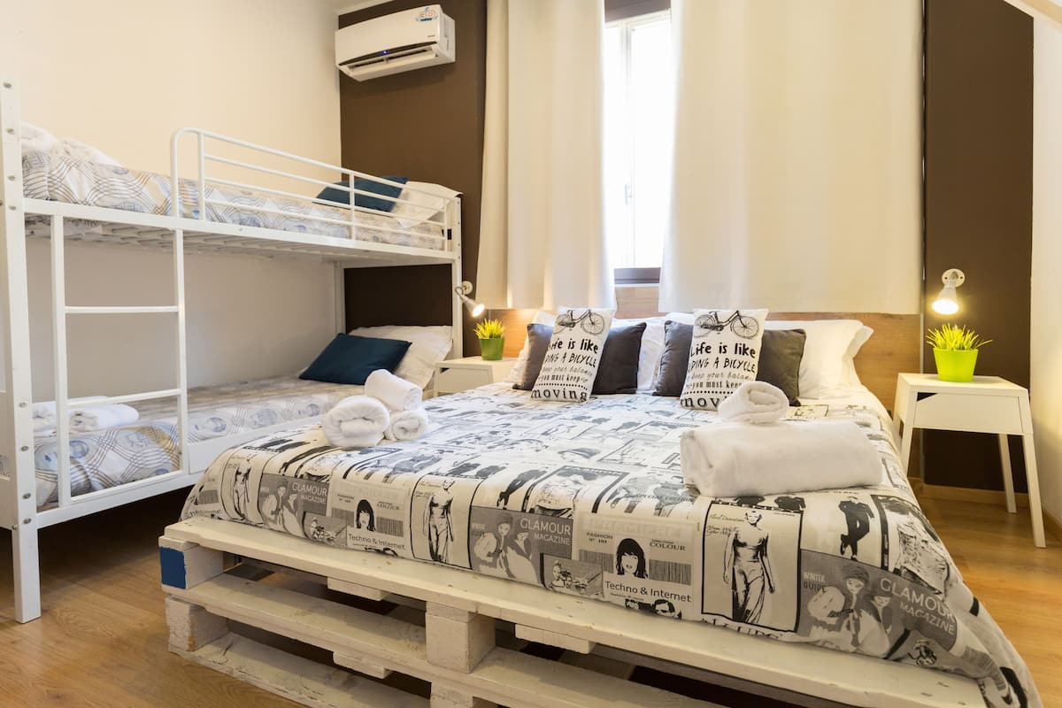 A room with bunk beds, a bed with patterned bedding, bedside tables with lamps, and a window with curtains.