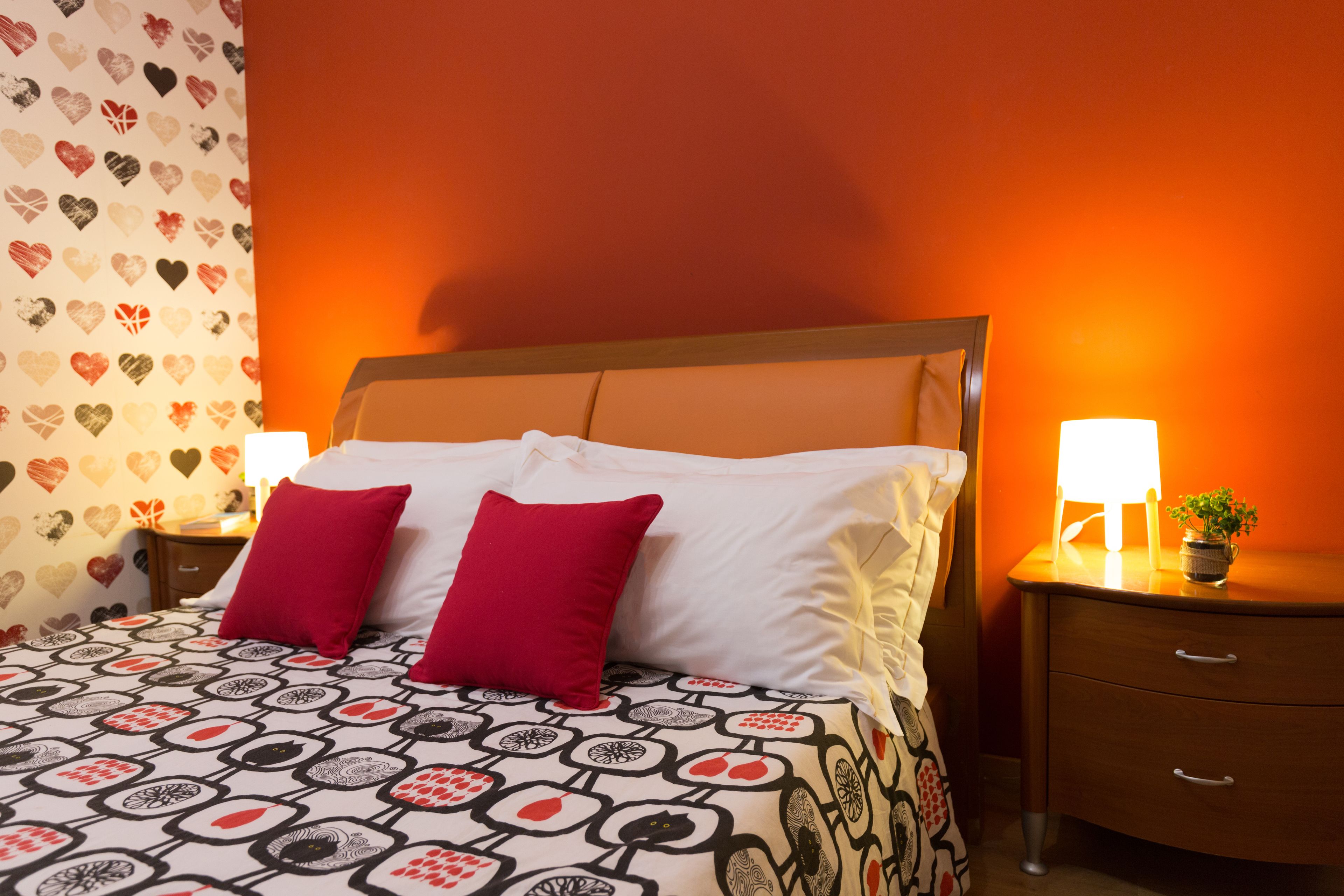 A neatly made bed with a patterned bedspread and two pillows, a nightstand with a lamp, and a wall with heart-patterned wallpaper.