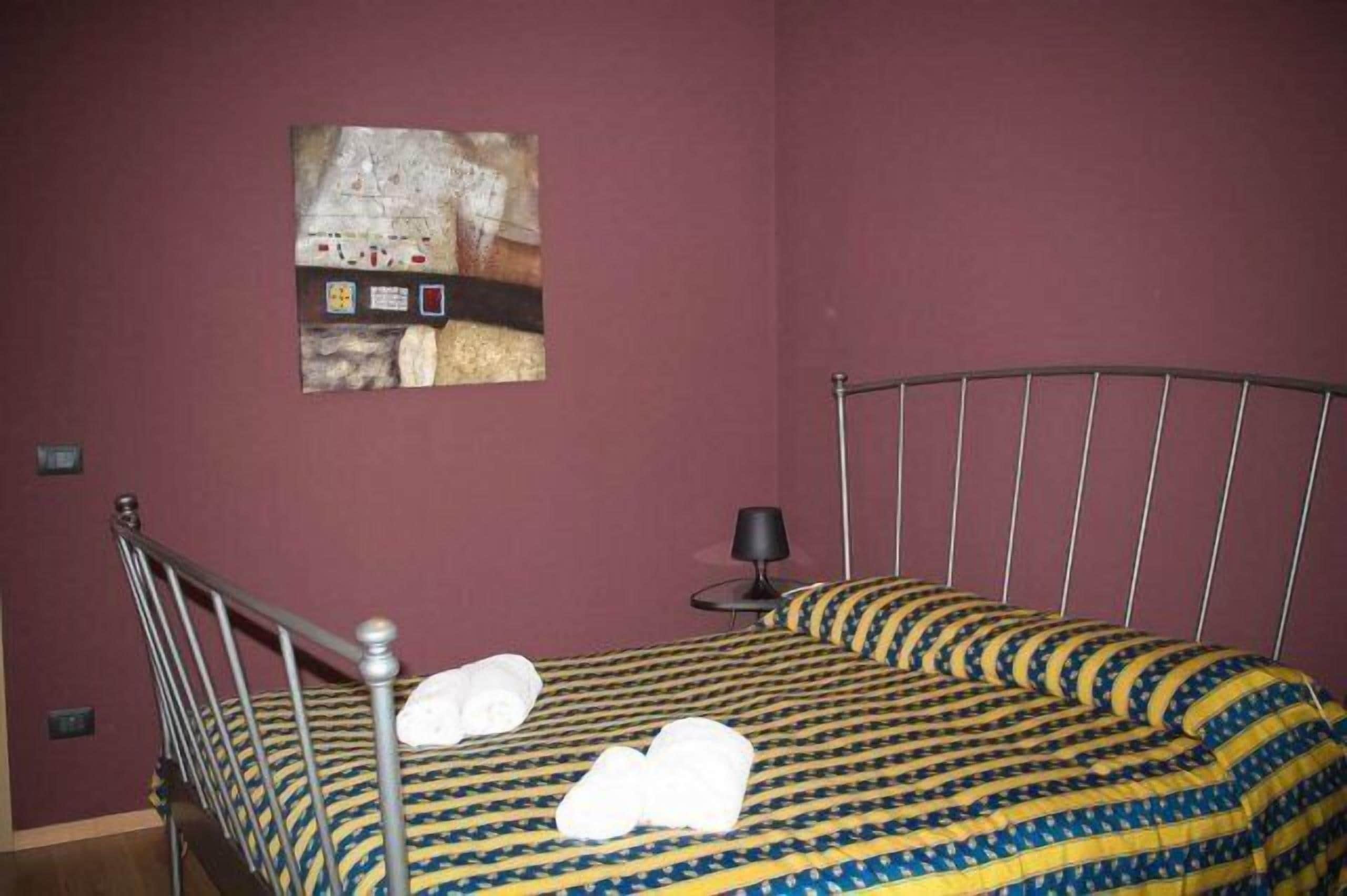 Una cama individual con estructura metálica, un edredón estampado y dos toallas enrolladas sobre la cama.
