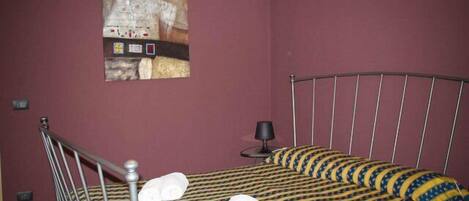 Una cama individual con estructura metálica, un edredón estampado y dos toallas enrolladas sobre la cama.