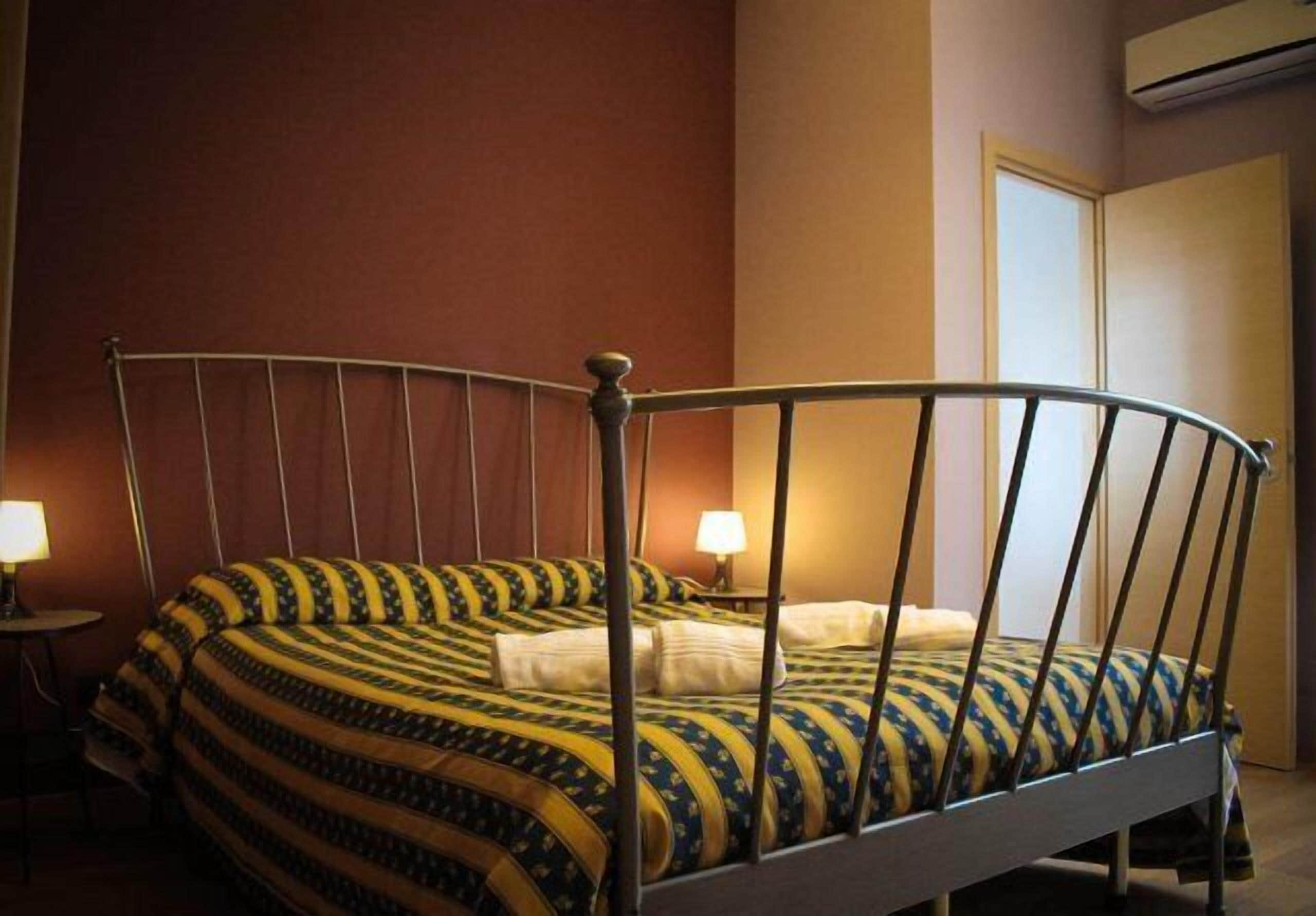 A single bed with a metal frame, a patterned bedspread, two bedside lamps, and a closed door in the background.