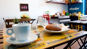 Una mesa con un pastel, una taza y un platillo. Hay sillas alrededor de la mesa y una estantería con diversos objetos encima.