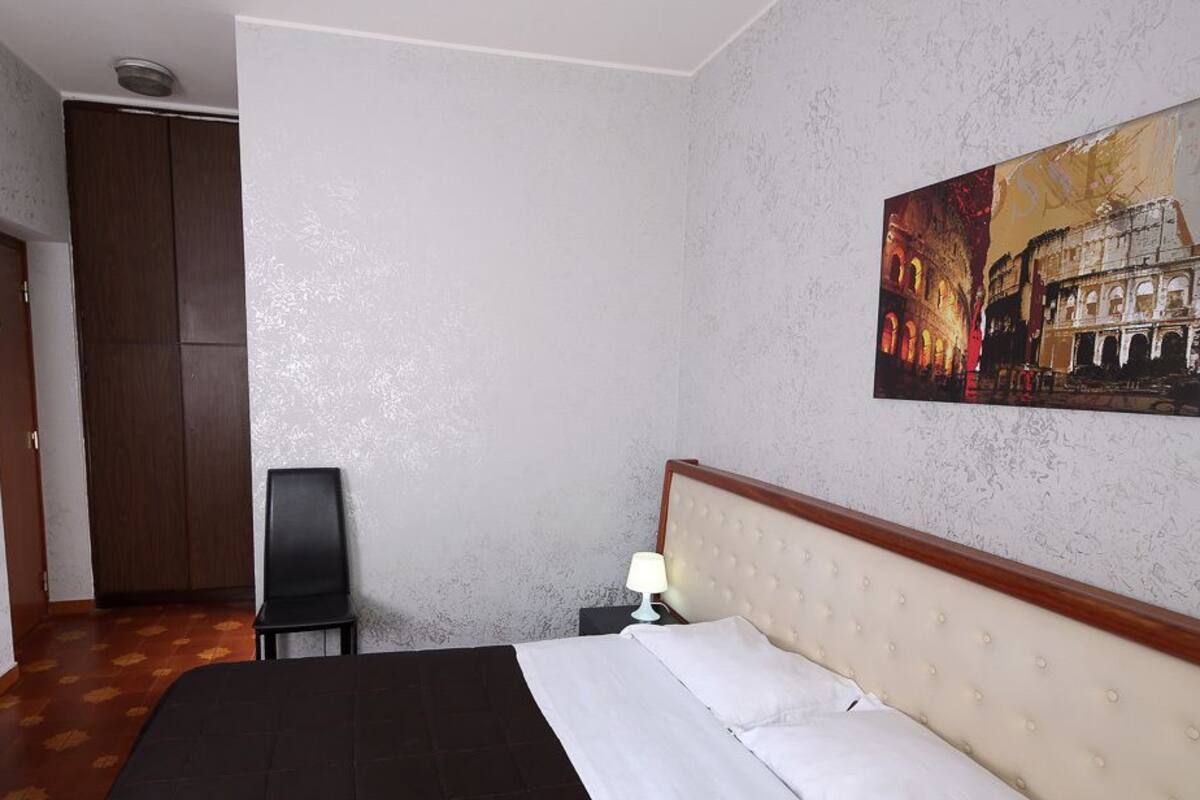 Une chambre d’hôtel comprenant un lit, un bureau, une chaise, un tableau représentant le Colisée et une armoire.