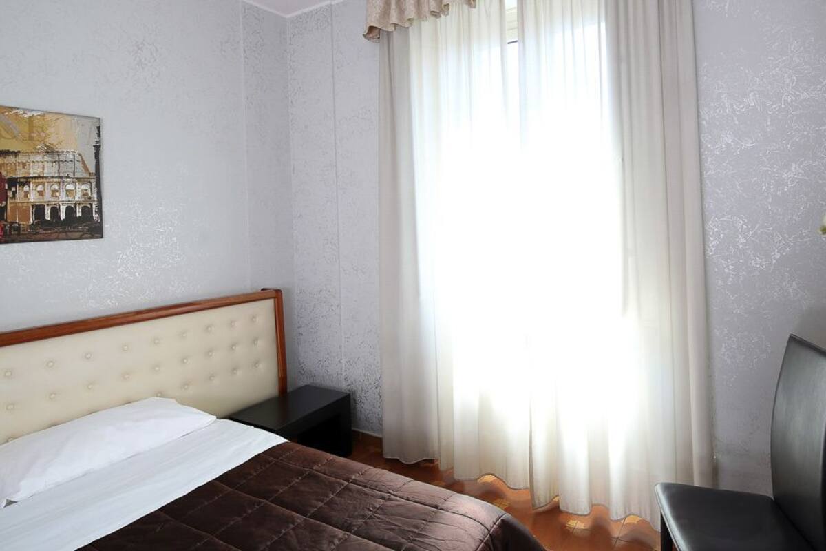 Une chambre à coucher comprenant un lit, une chaise, une table de chevet, une fenêtre avec des rideaux légers et un tableau accroché au mur.