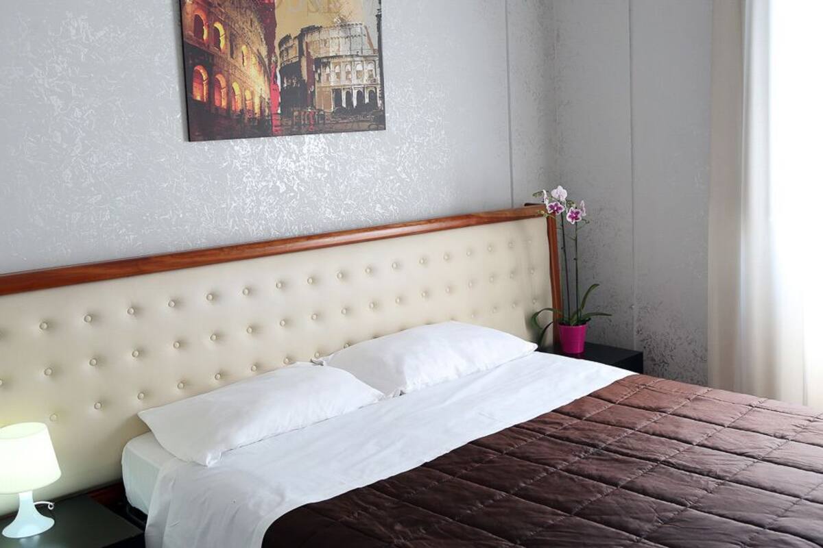 Un lit bien fait, avec une couette blanche et une couverture marron, une tête de lit en bois, une lampe de chevet et un pot d’orchidée.