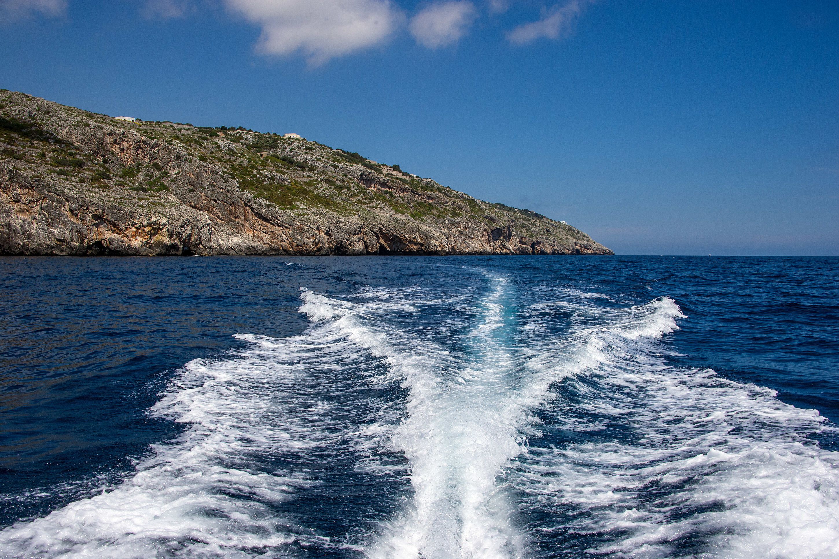 La estela de un barco en el océano, con una costa rocosa y un cielo despejado.