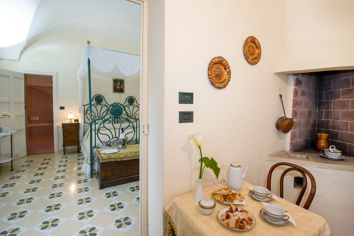 Una habitación con una cama, una mesa dispuesta con platos, tazas y un florero con una flor. Hay una chimenea y una pared adornada con placas decorativas.