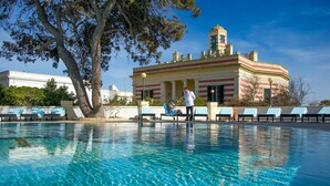 Piscina com espreguiçadeiras e um edifício com colunas ao fundo.