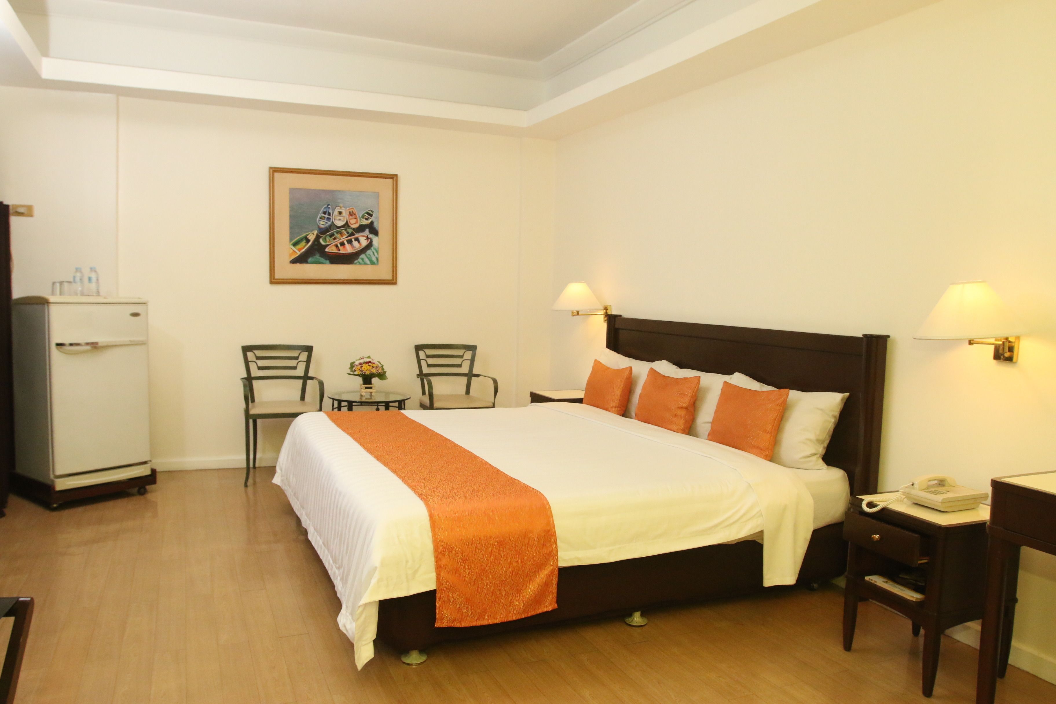 A hotel room with a large bed, two bedside tables with lamps, a desk with a telephone, a chair, a small table with a vase, and a refrigerator.