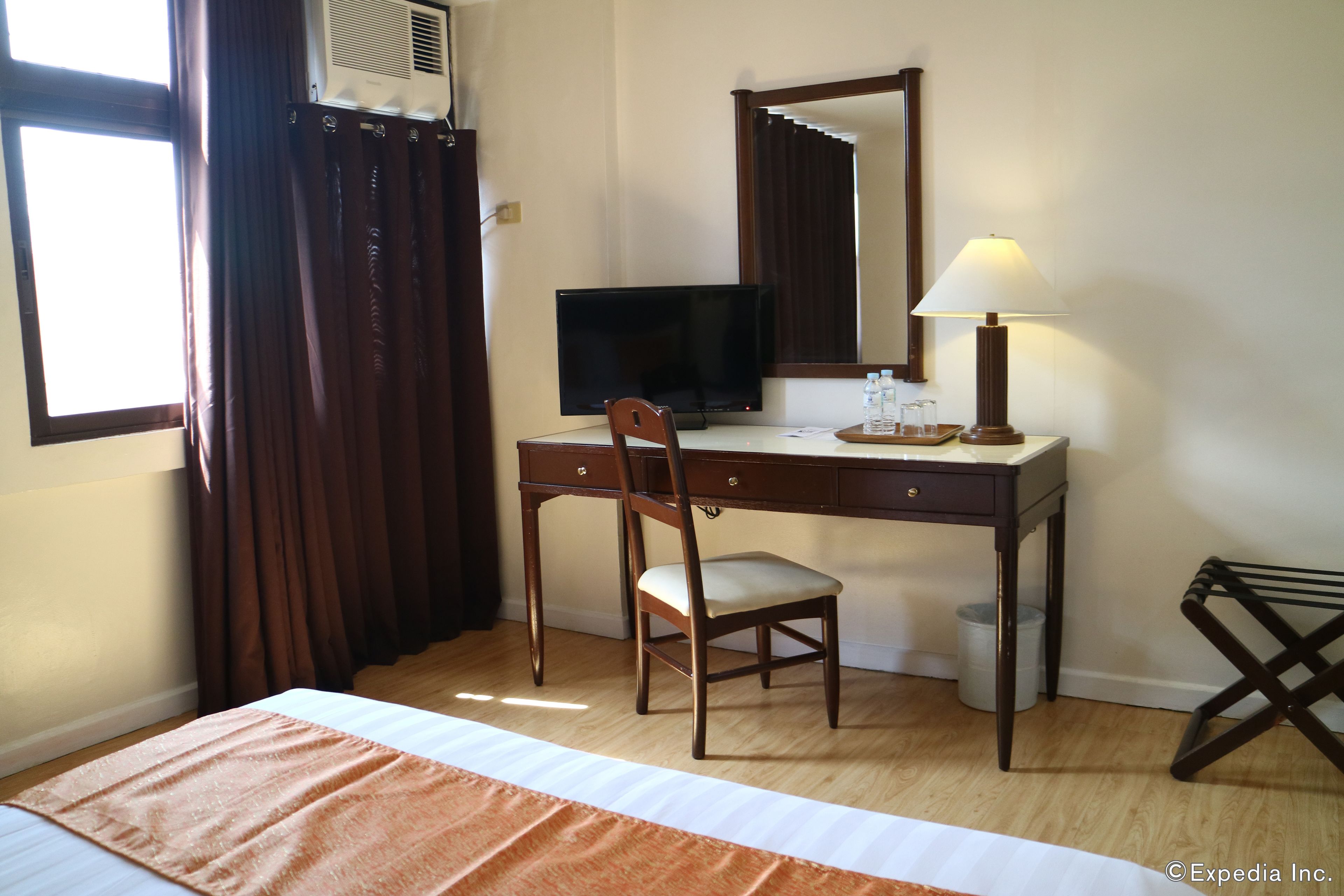 Una habitación de hotel con cama, escritorio, silla, televisor, lámpara y ventana con cortinas.