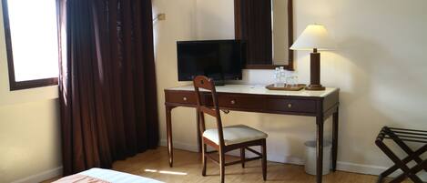 Una habitación de hotel con cama, escritorio, silla, televisor, lámpara y ventana con cortinas.
