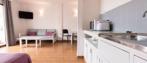 A modern kitchen with a stainless steel sink, a microwave, and a white cabinet. There is a small dining table with a chair and a sofa with cushions.