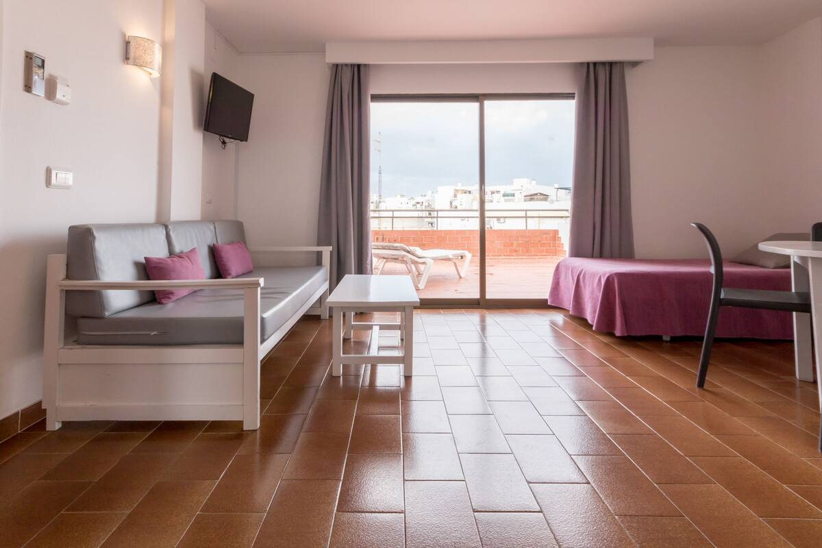 Una habitación de hotel moderna con balcón, un sofá, una mesita y una cama con colcha morada.