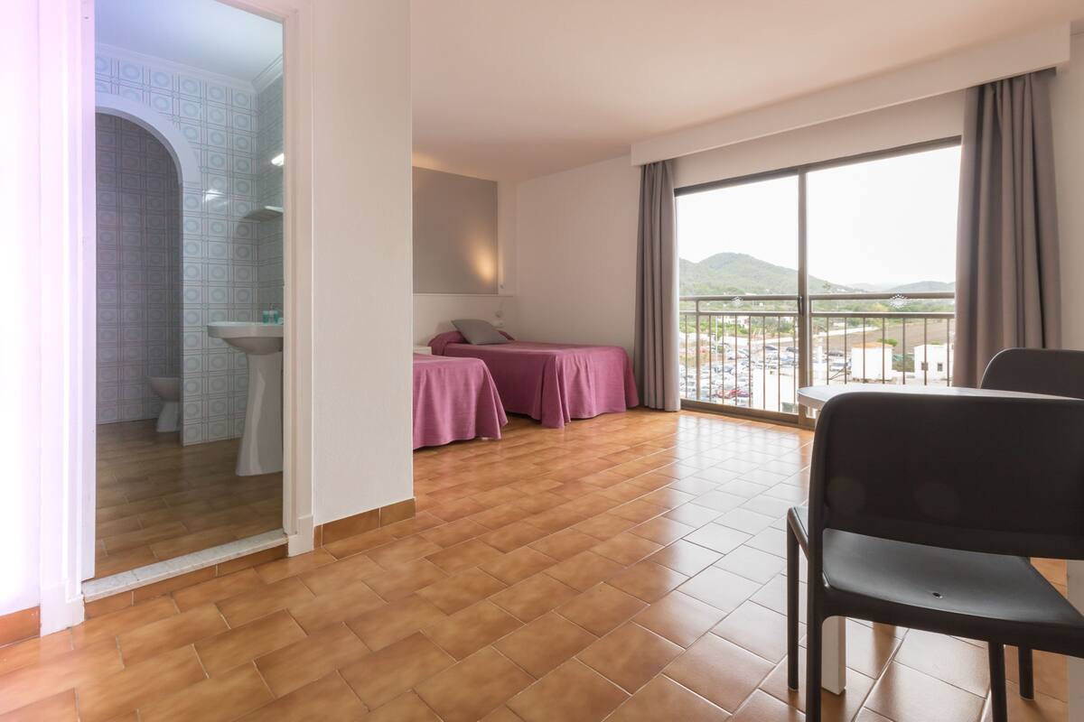 Habitación de hotel con piso de madera, balcón con vistas y baño con pared de azulejos. Incluye cama con cubrecama rosa.
