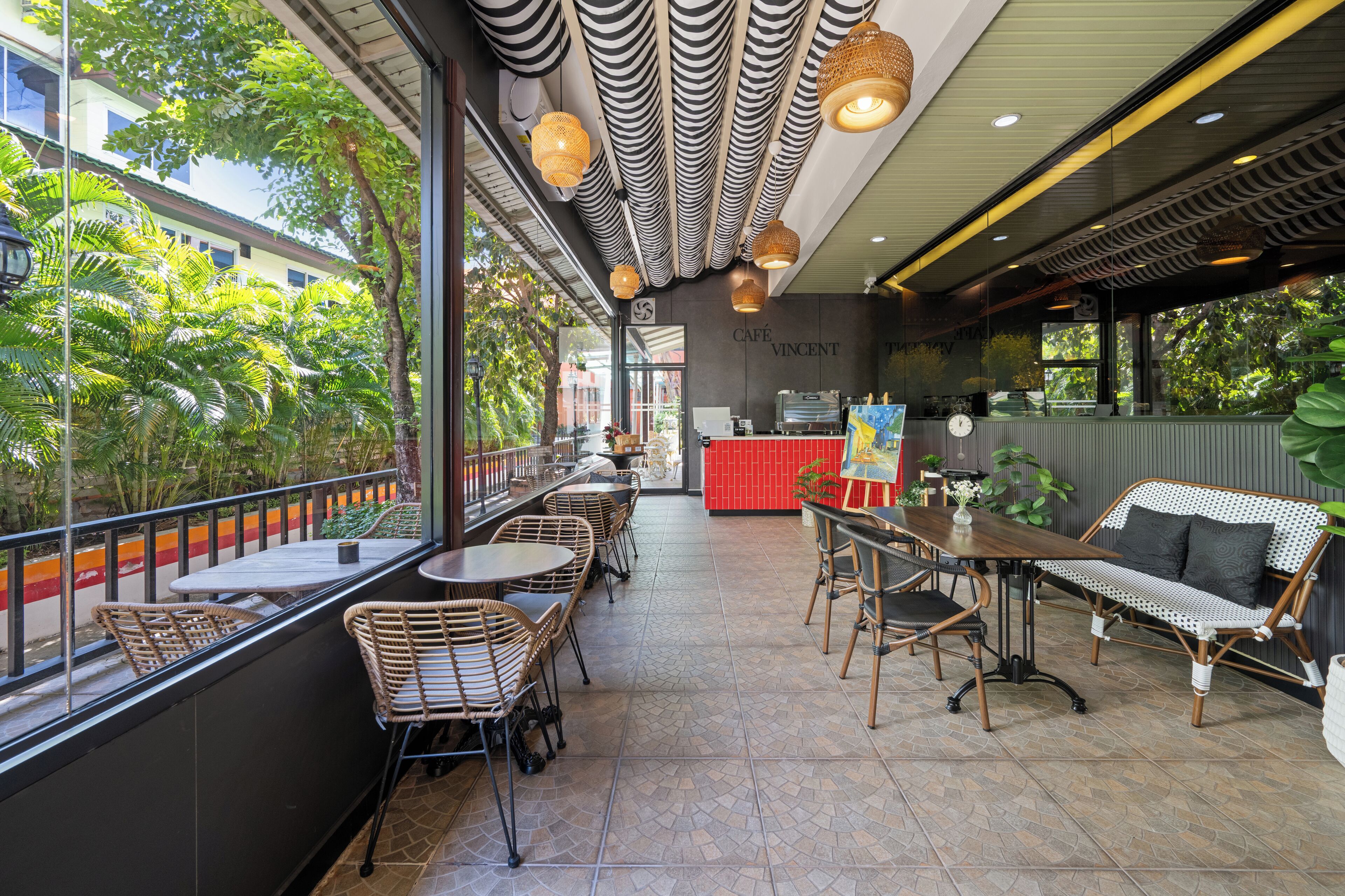 A café with wicker furniture, a red counter, and large windows overlooking greenery.