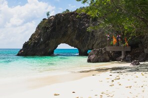 Une arche naturelle formée par la roche, agrémentée d’un panneau en bois et de drapeaux, qui surplombe une plage de sable et des eaux turquoise.